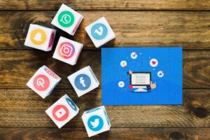 Colorful social media logo dice scattered around a blue puzzle featuring a computer screen and engagement icons on a wooden table.