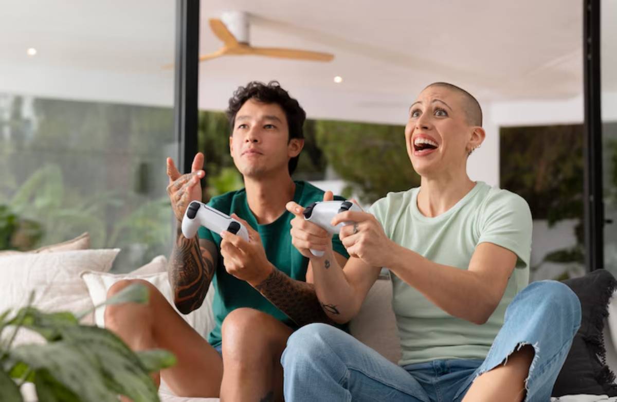 Two friends are sitting on a couch, intensely focused on gaming with white controllers, in a modern, sunlit living room.